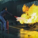 A woman uses a fire extinguisher to put out a large flame in an outdoor setting, showcasing fire safety.